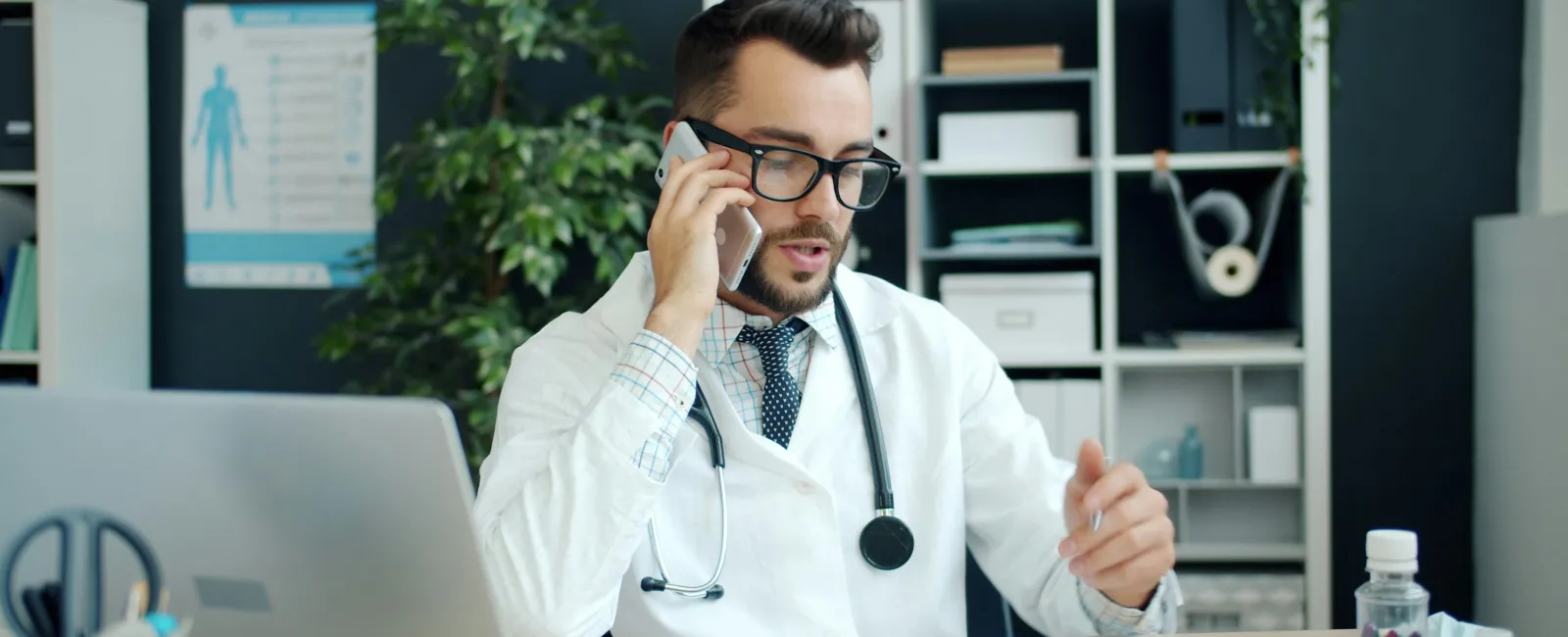 Male doctor in white coat with stethoscope on phone call at desk with medical documents and laptop.