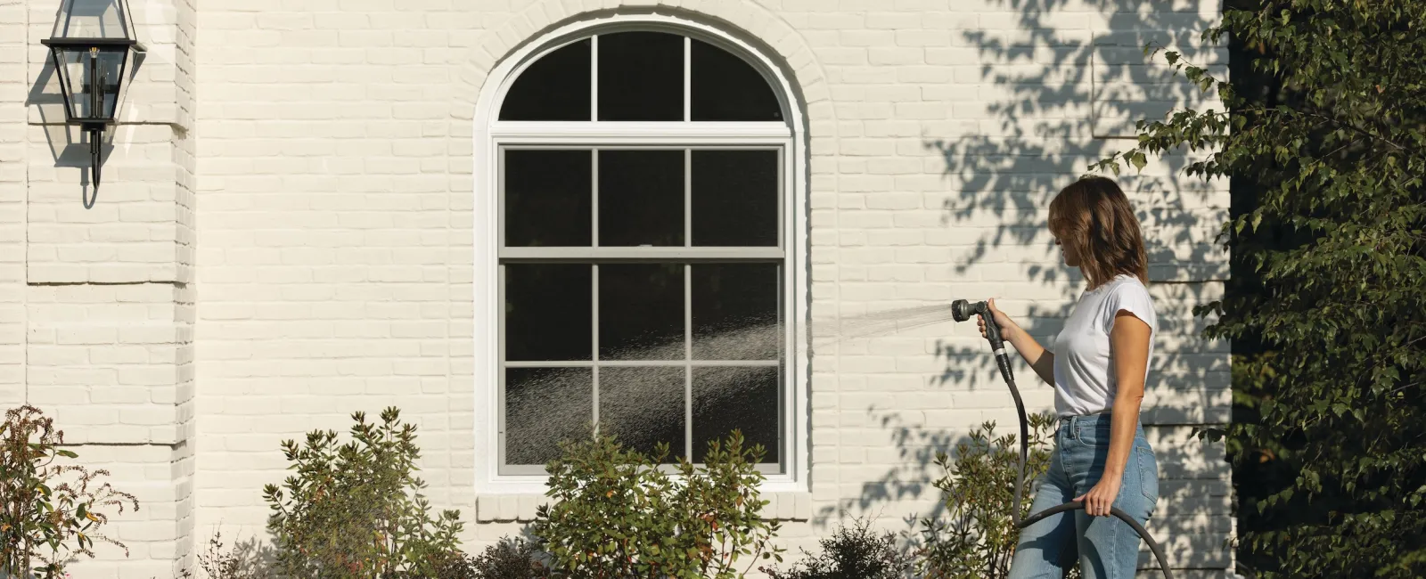 Woman watering a flower garden with a hose in front of a white brick house with an arched window.