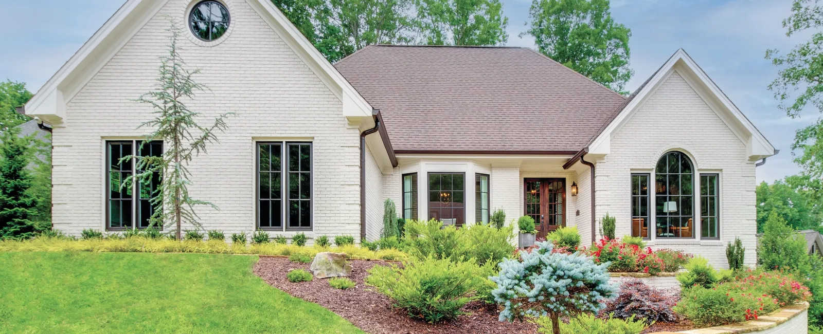 Modern white brick house with gable roof, large windows, and landscaped front yard with shrubs and trees.