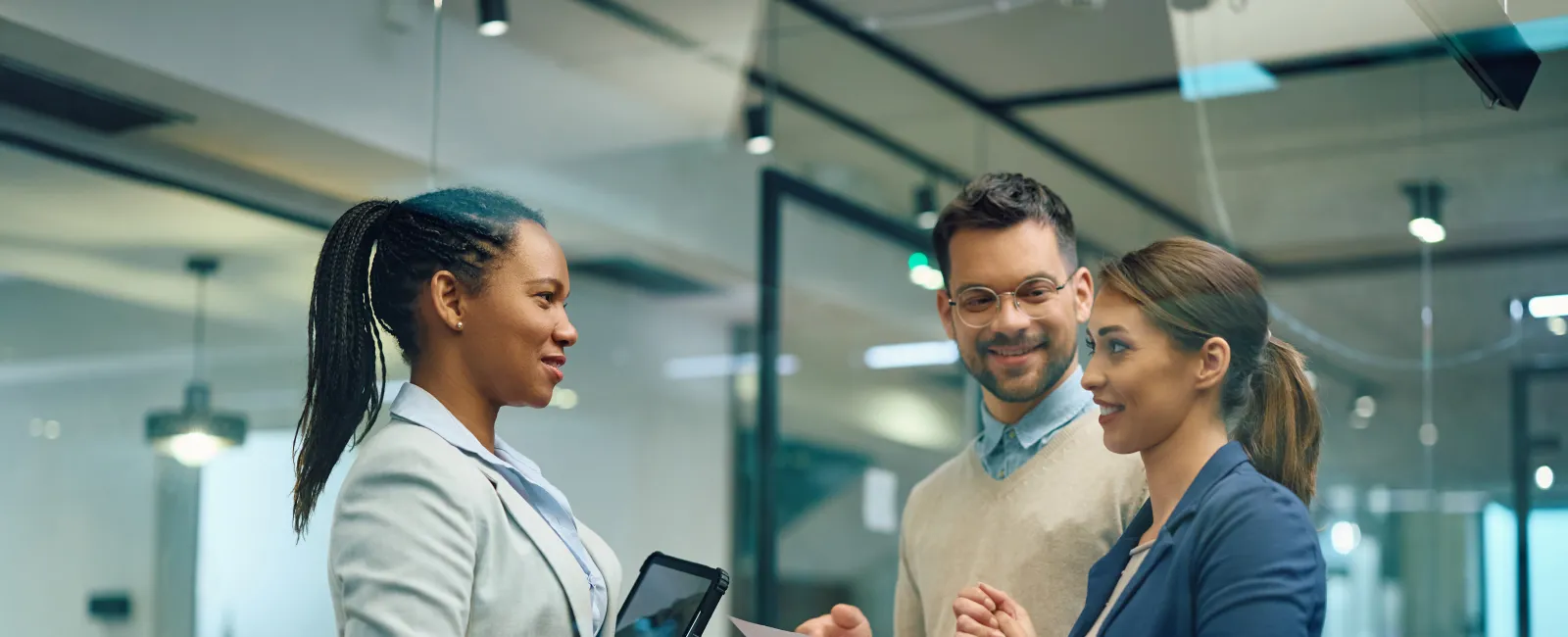 Three professionals engage in a discussion inside a modern office with papers and a tablet.