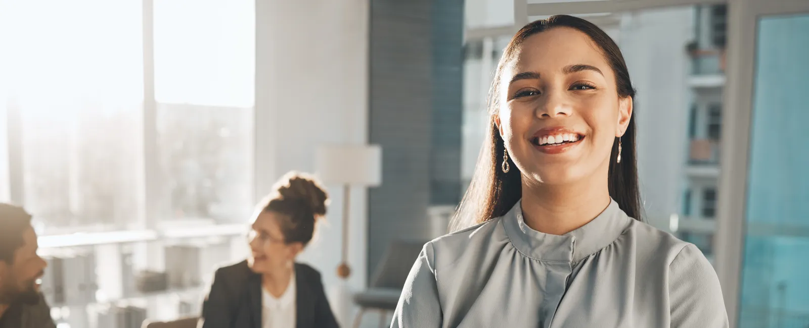 Confident young woman standing with crossed arms in modern office with colleagues talking in background