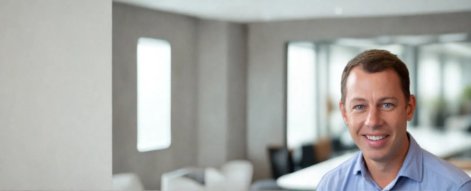 Smiling man in blue shirt sitting in a bright modern office with large windows and blurred background.