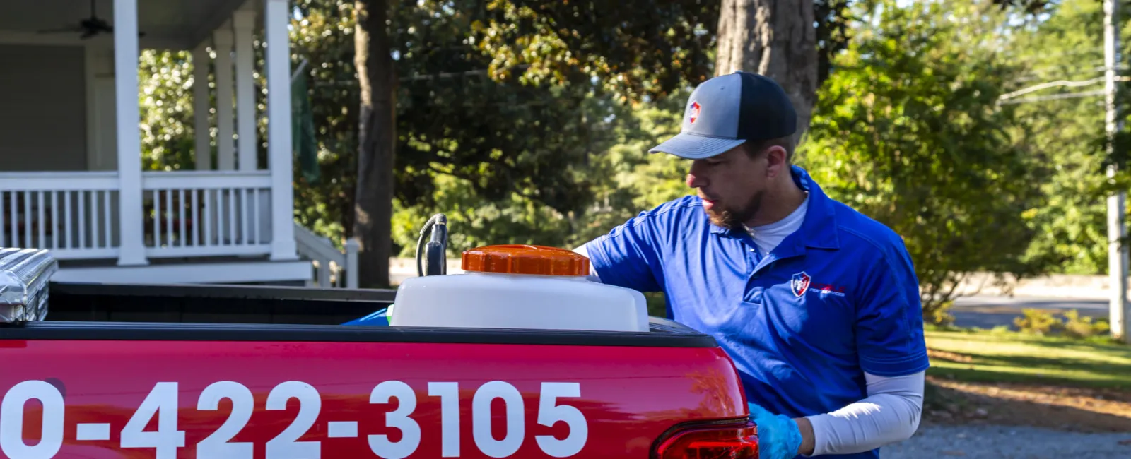 Pest control technician in blue uniform working at rear of red service truck with phone number on side.