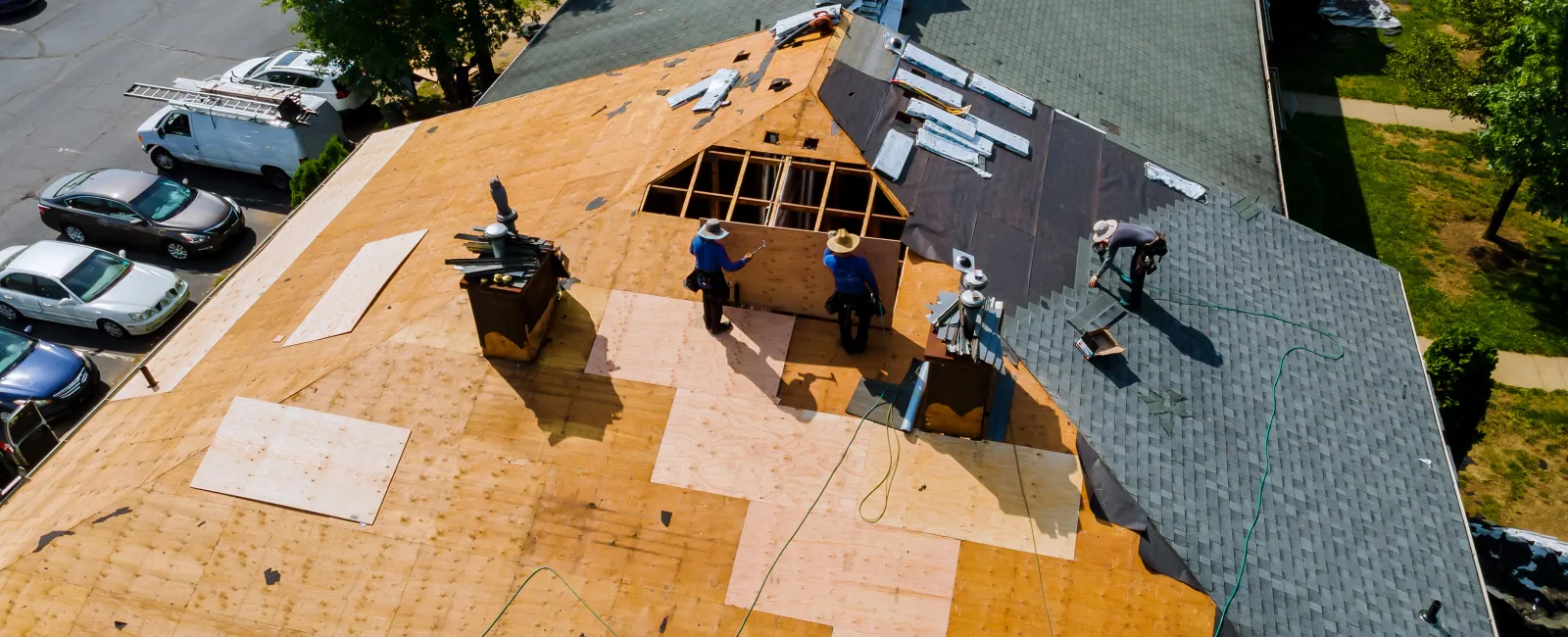 Workers installing new roof plywood and shingles on large building roof during daytime in sunny weather