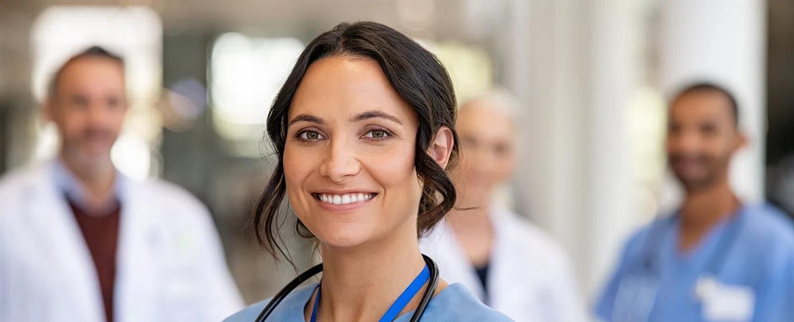 Smiling female nurse in blue scrubs with stethoscope, medical team blurred in background in hospital.