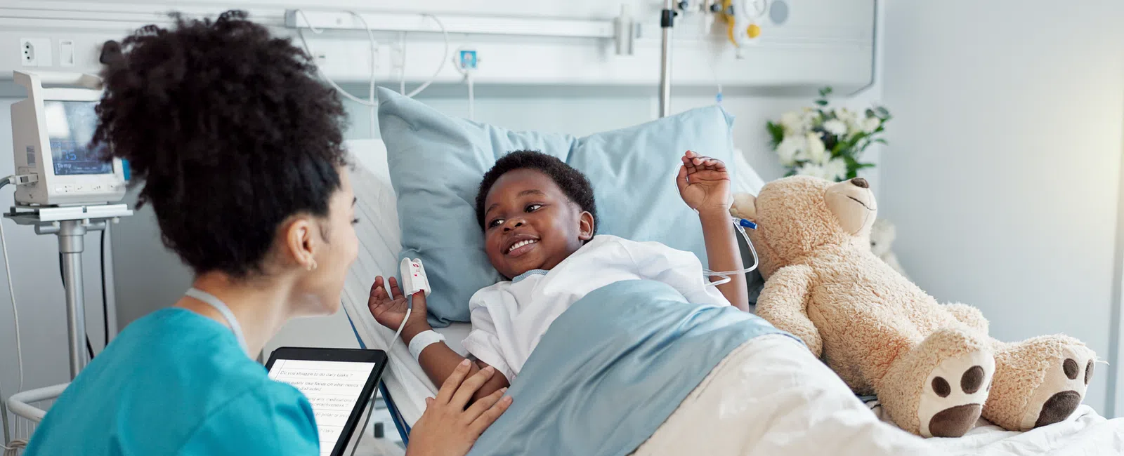 Smiling pediatric nurse interacts with happy child patient lying in hospital bed with teddy bear and medical equipment.