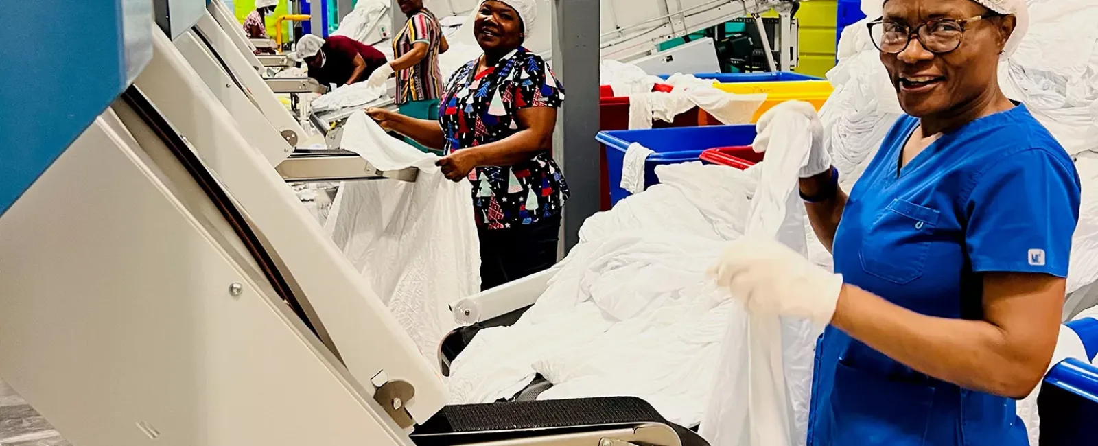 Workers in hairnets and gloves processing white linens using industrial machines in a textile factory.