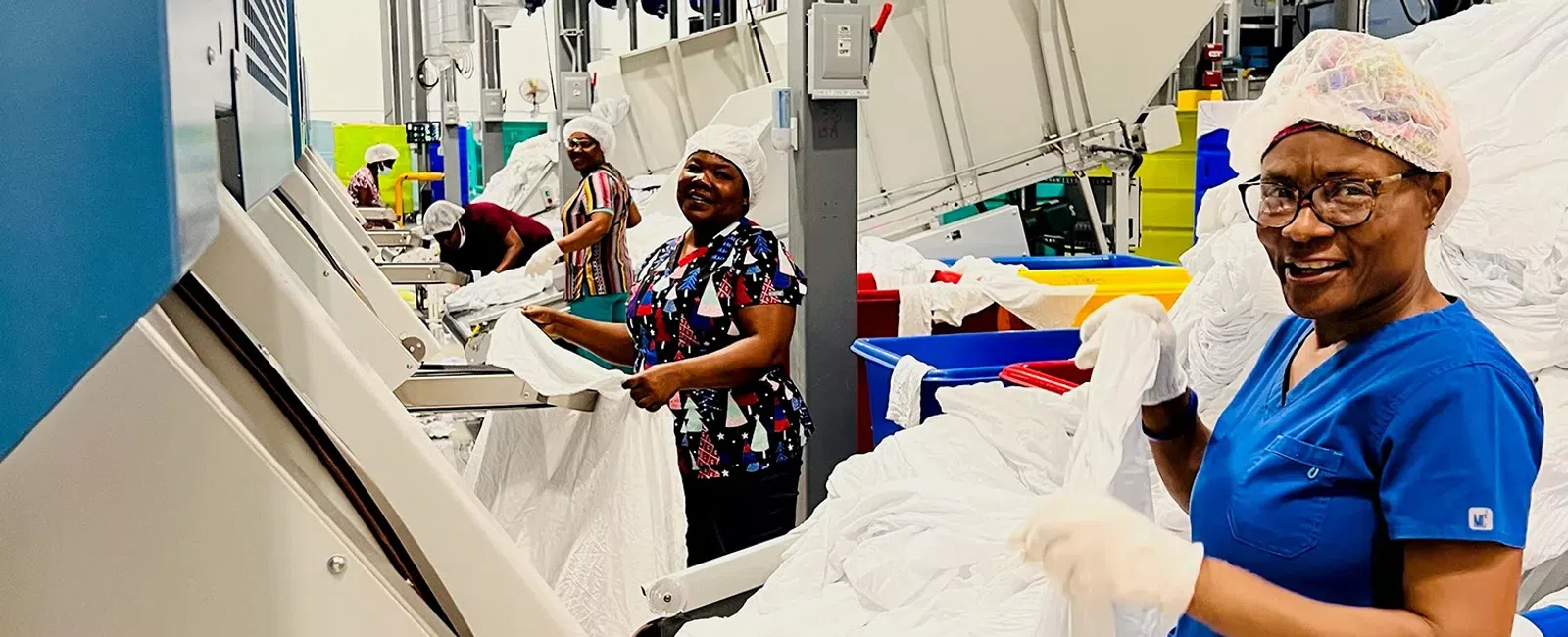 Workers in hairnets and gloves processing white linens using industrial machines in a textile factory.