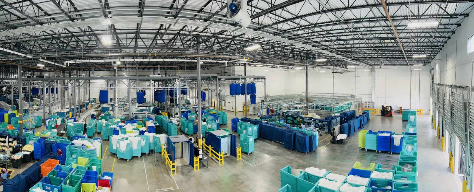 Wide view of a textile factory with machines, fabric bins, and high ceilings in a spacious industrial setting.