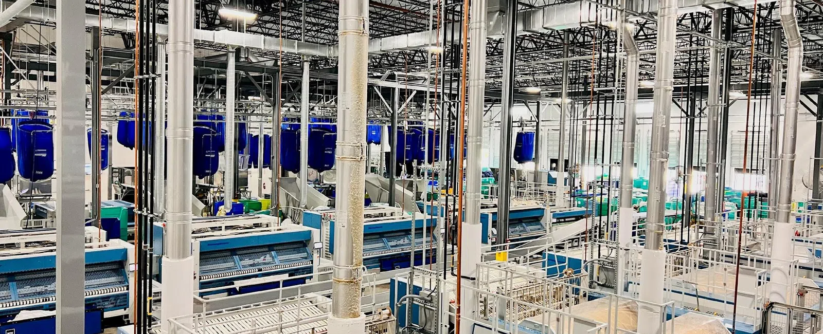 Large industrial textile factory interior with rows of blue machinery and metal piping under a high ceiling.