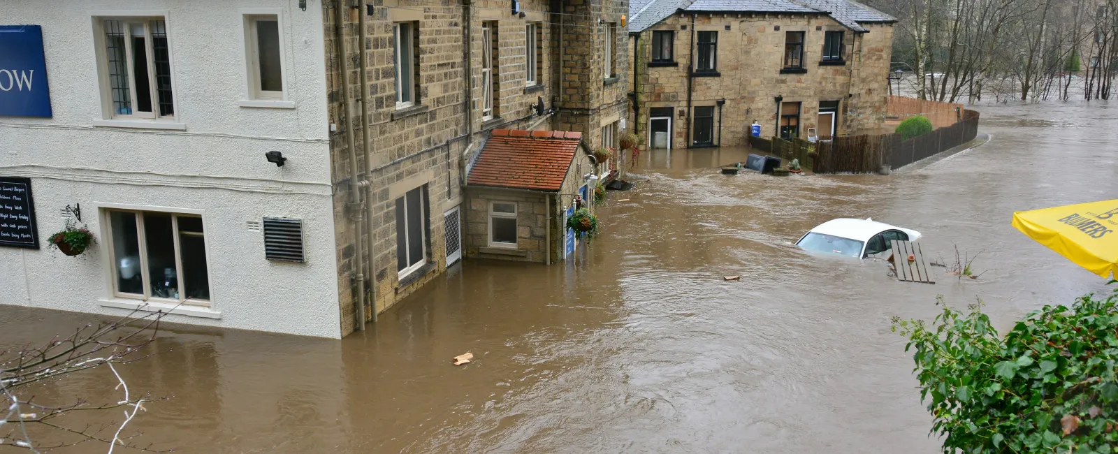 Floodwaters surround stone houses and partially submerge cars on a residential street during heavy flooding.