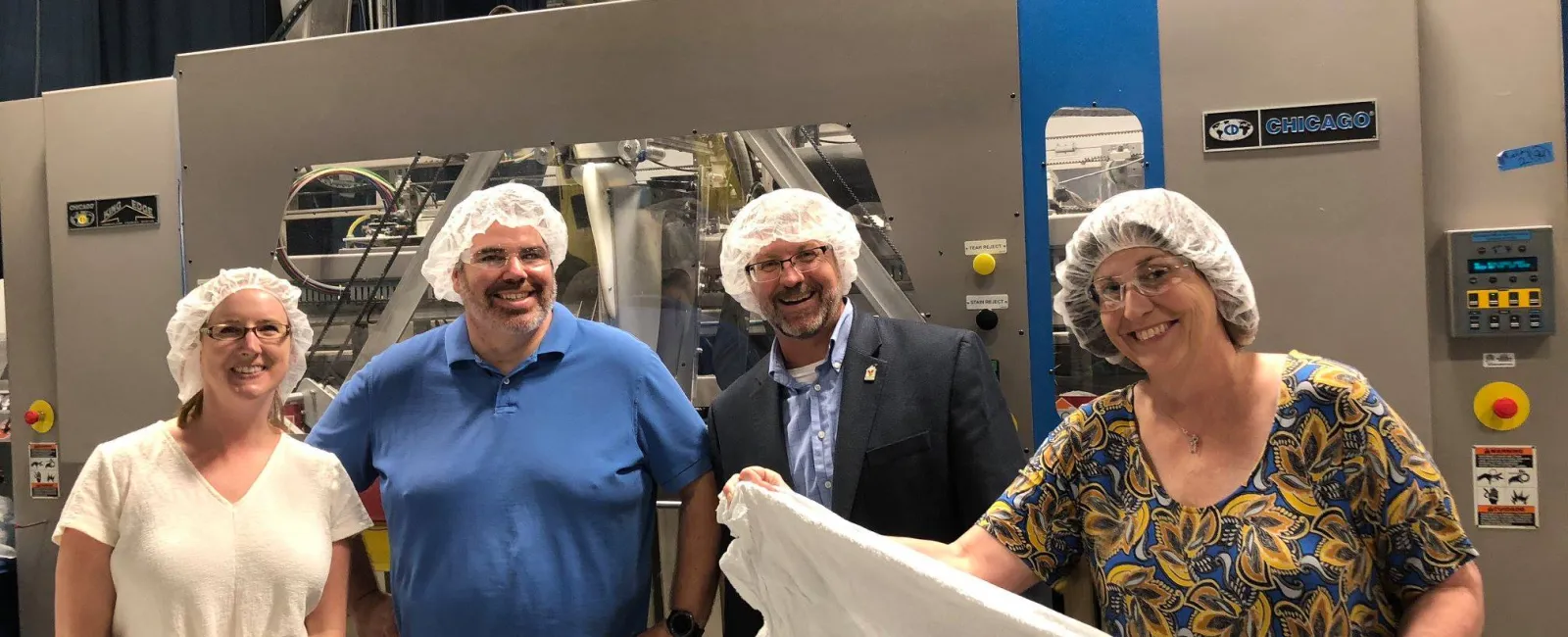 Four people wearing hairnets stand in a factory, one holding a large white sheet of material near machinery.