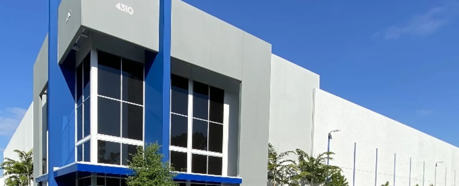 Modern commercial building with blue accents and large windows under a clear blue sky, featuring parking and greenery