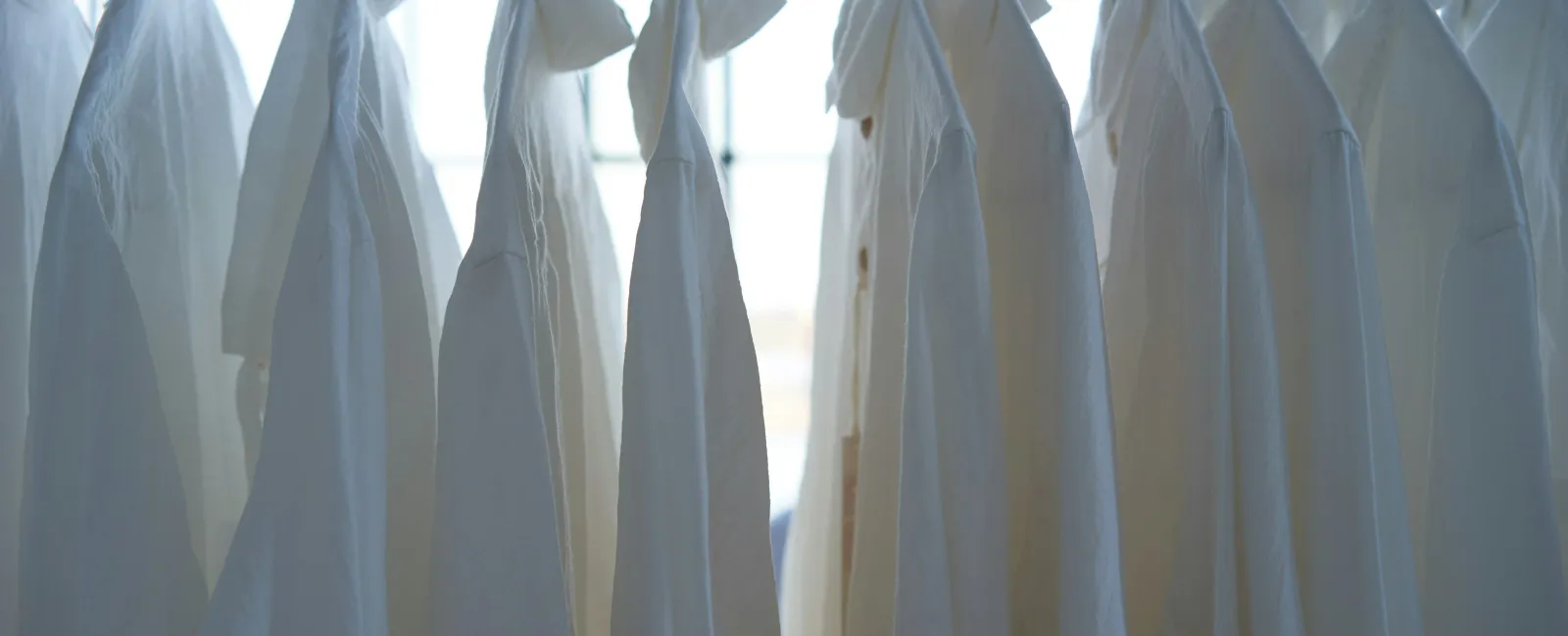 White dress shirts hanging in a row on hangers in front of a bright window.