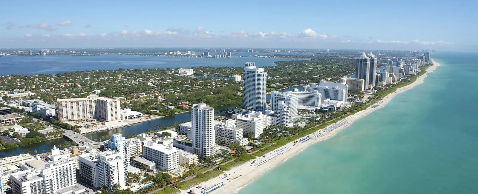 Aerial view of Miami Beach coastline with turquoise ocean, sandy beach, and high-rise buildings under clear blue sky.