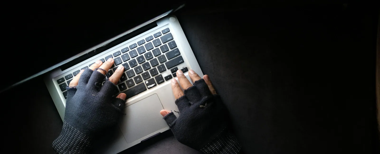 Hands in fingerless gloves typing on a laptop keyboard over a dark surface with moody lighting