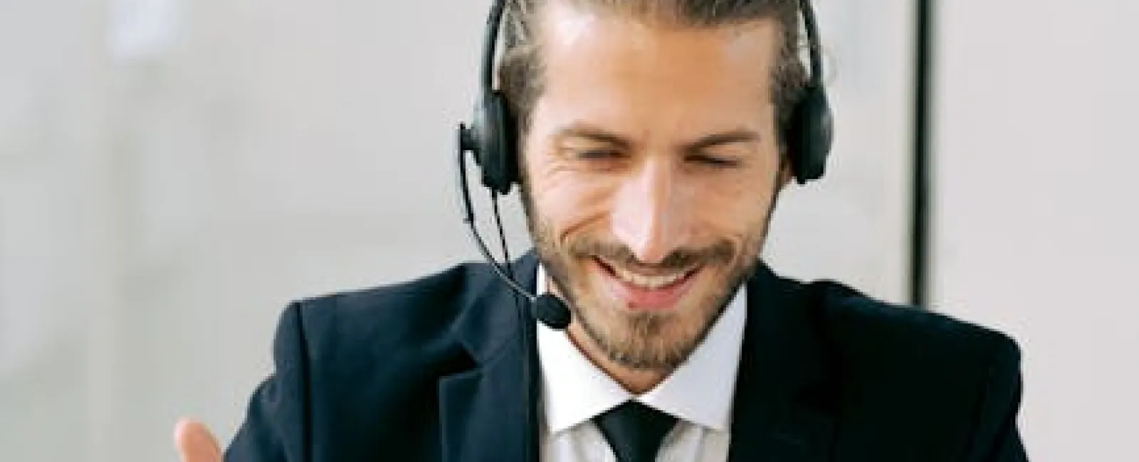 Smiling man in suit with headset working on laptop and gesturing during video call in office