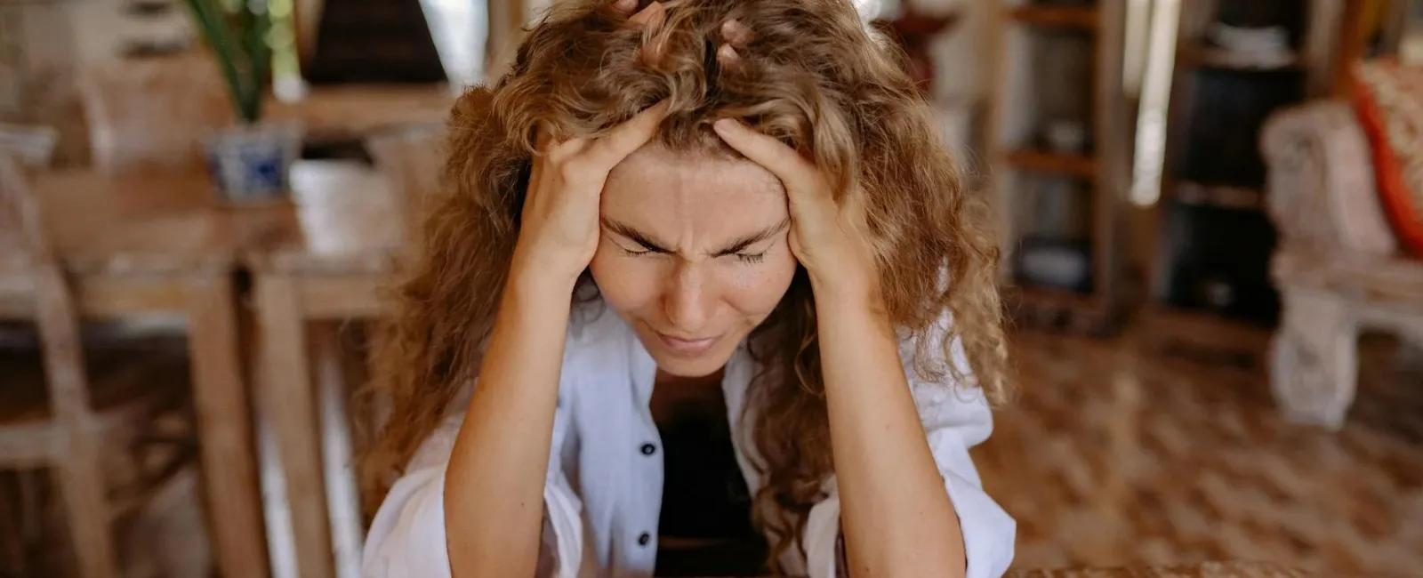 Frustrated woman sitting at wooden table holding head with hands in front of laptop and smartphone.