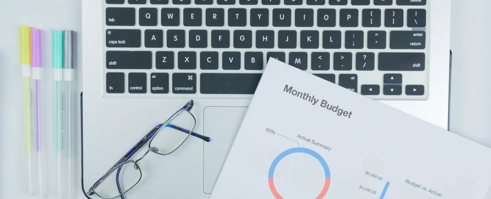 Glasses, pastel highlighters, and a monthly budget report on a laptop keyboard workspace.