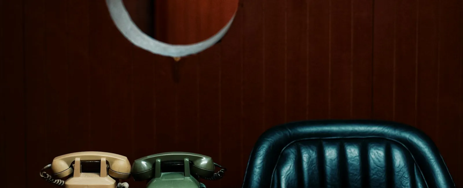 Retro beige and green rotary phones on a dark table with a blue leather chair and brown wooden wall background.