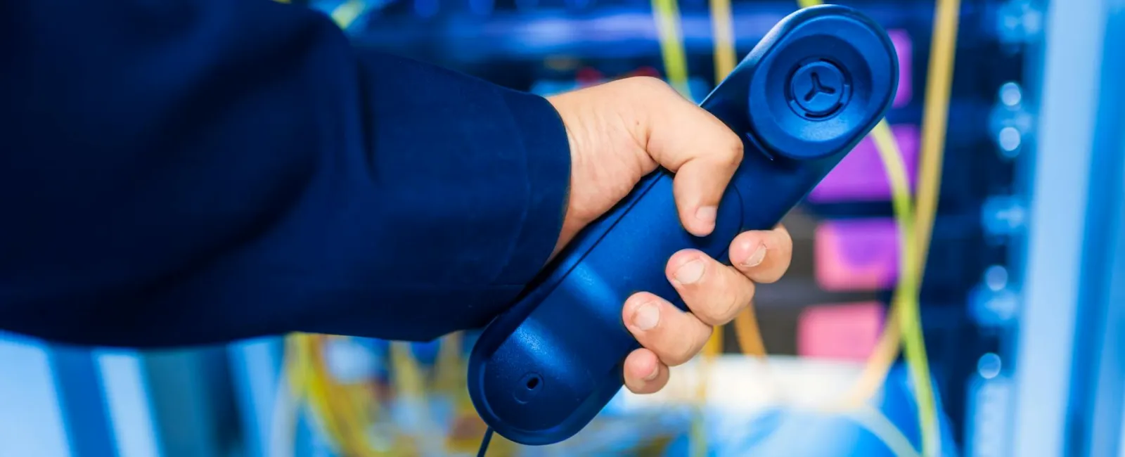 Person holding a blue telephone handset with network cables and server hardware in the background.