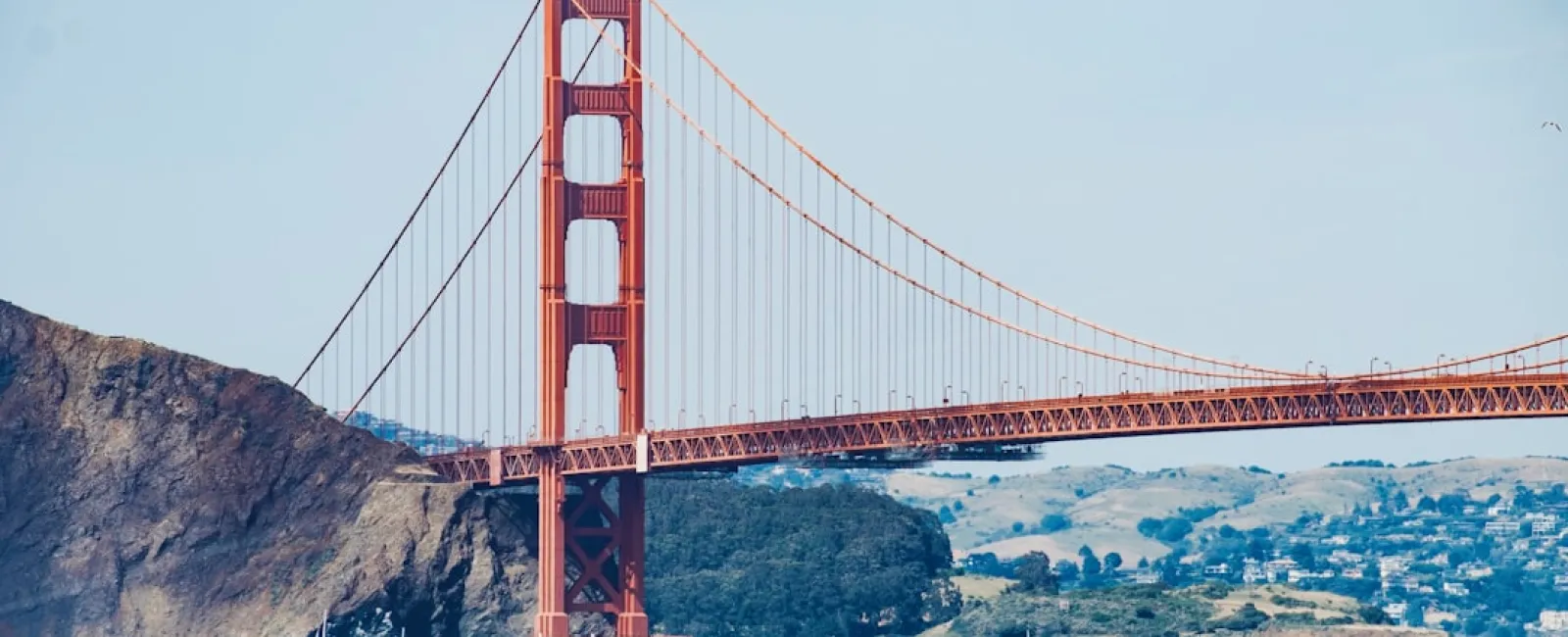 Golden Gate Bridge spans across blue water with rocky hills and clear sky in the background under daylight.