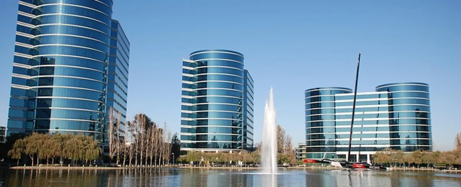 Modern office buildings with reflective glass facades beside a lake with a central fountain under a clear blue sky