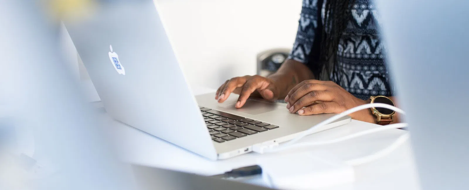 Person typing on a silver MacBook laptop at a white desk with charging cables connected.