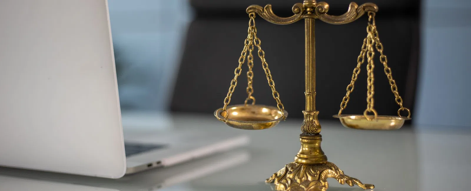 Golden balance scale on glass desk next to open laptop in modern office setting with blurred background.
