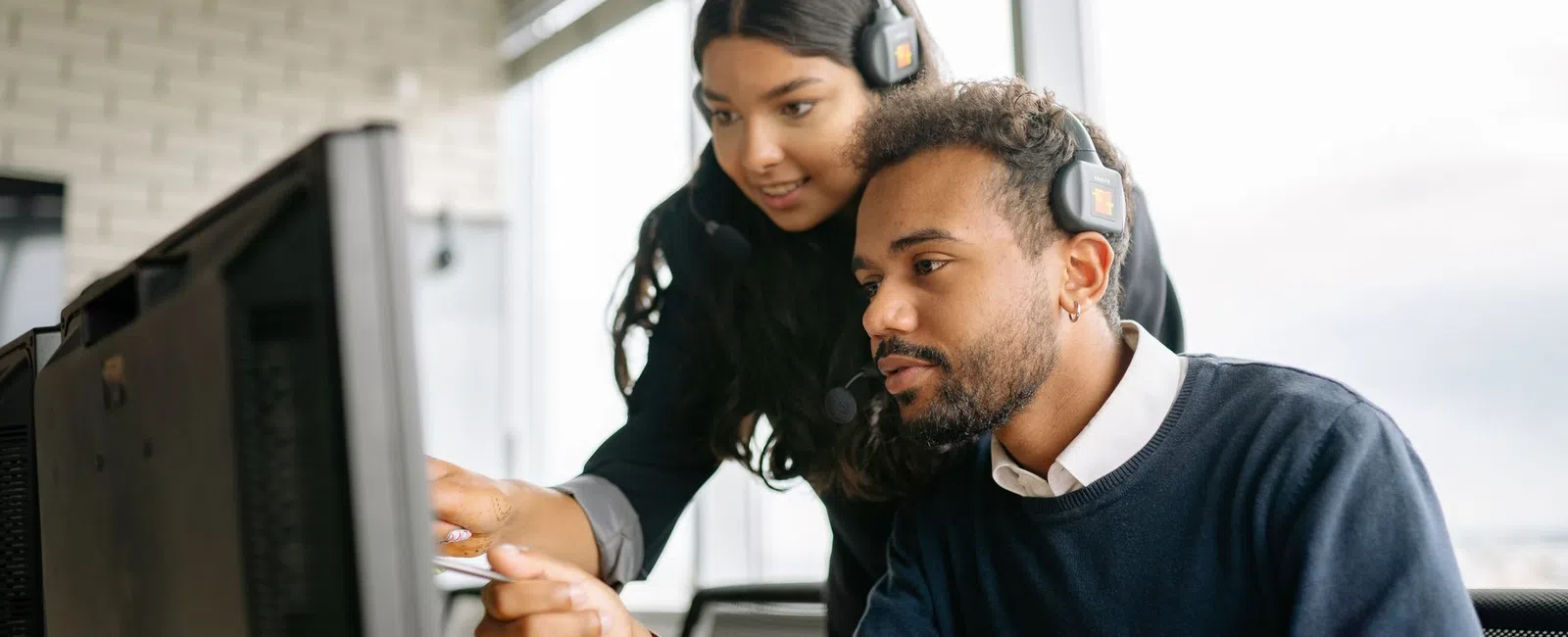 Two colleagues wearing headsets focused on a computer screen in a modern office workspace.