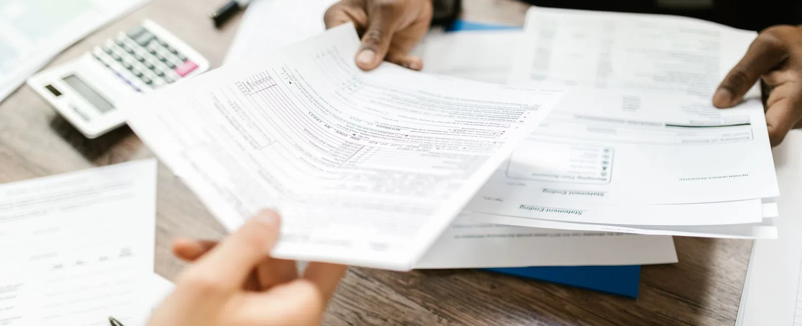 Two people exchanging financial documents over a wooden desk with calculator and paperwork nearby