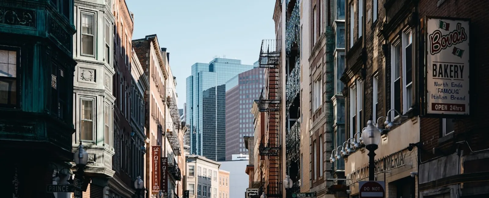Historic urban street lined with brick buildings and fire escapes leading to modern skyscrapers in the background