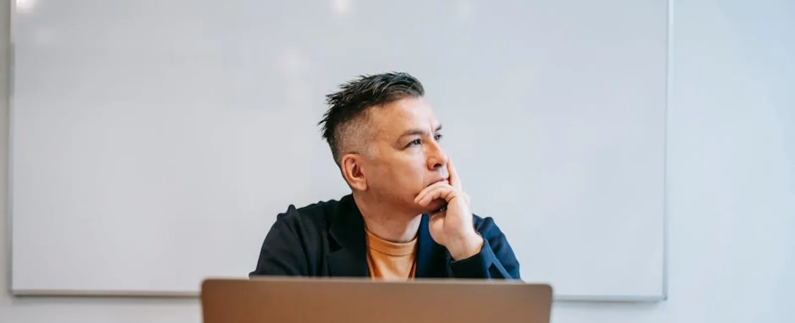 Man in office thinking deeply while sitting at desk with laptop, papers, and folders against a whiteboard