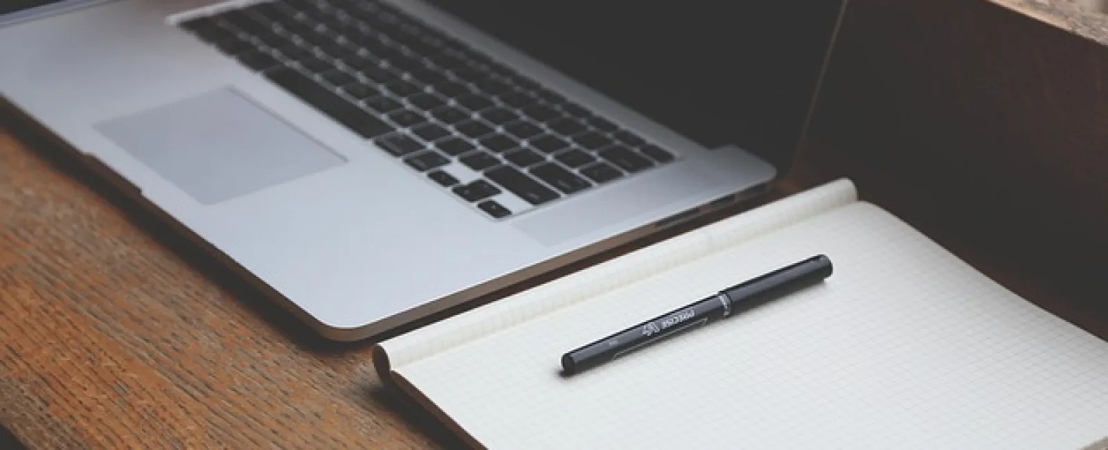 Open laptop and blank graph paper notebook with pen on a wooden desk for work or study.