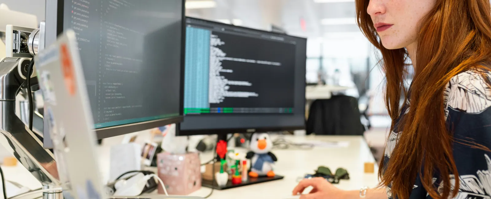 Female cyber security specialist working at a desk with dual monitors displaying code in a modern office.
