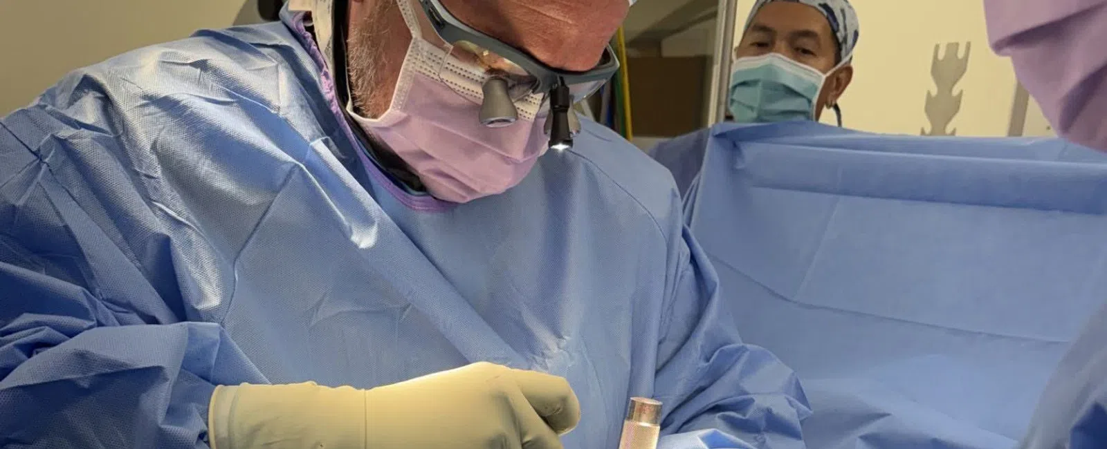 Surgeon wearing magnifying glasses and protective gear performing surgery in an operating room.