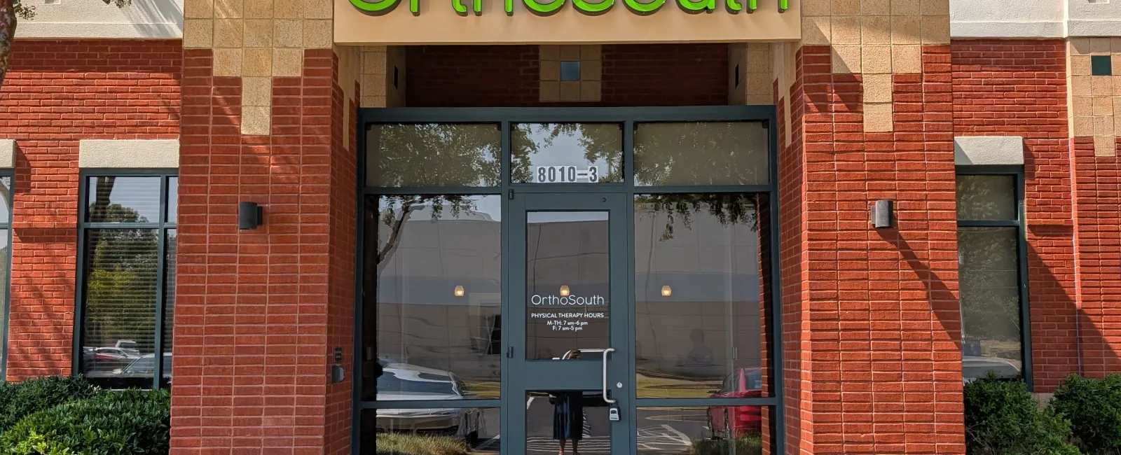 Entrance of OrthoSouth building with glass door, brick walls, greenery, and a curved black awning under a clear blue sky.
