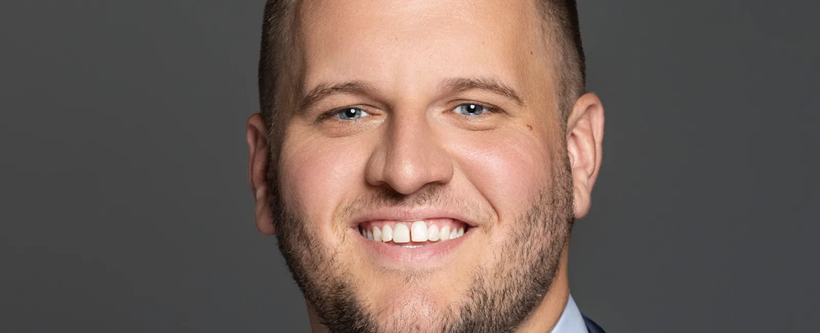 Smiling man with short hair wearing a navy suit, white shirt, and red striped tie against a gray background