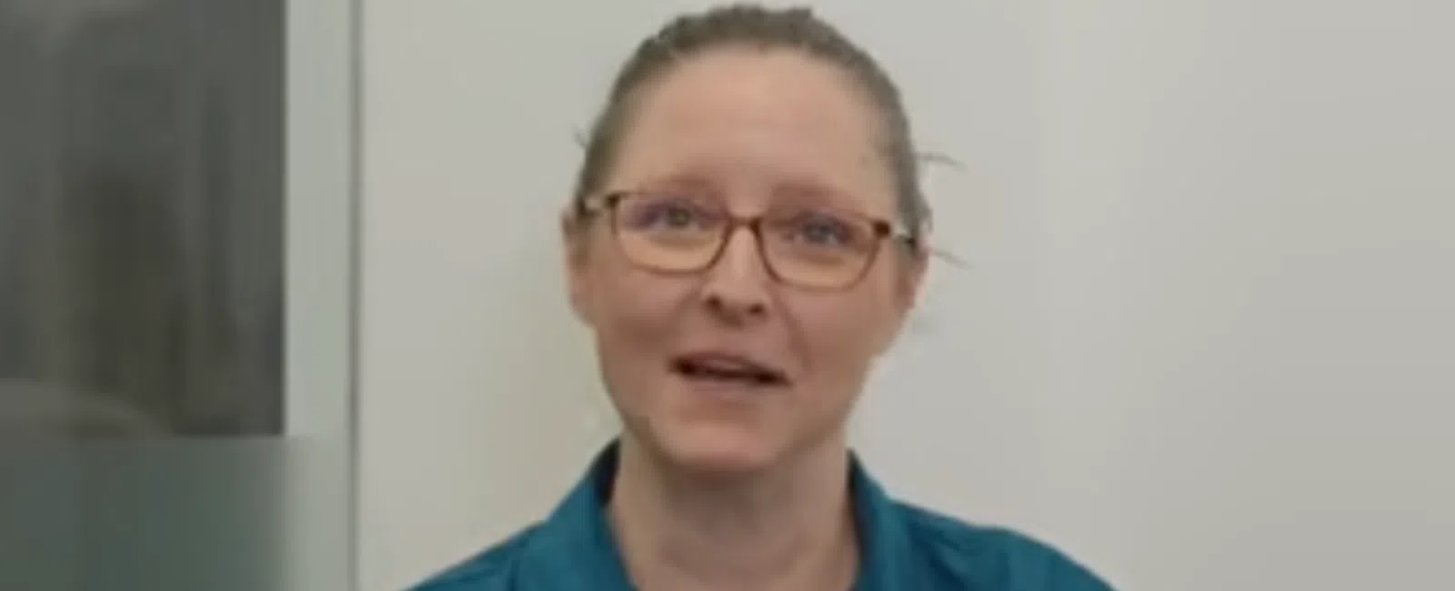Woman with glasses and teal shirt speaking inside a modern office with glass walls and white background.