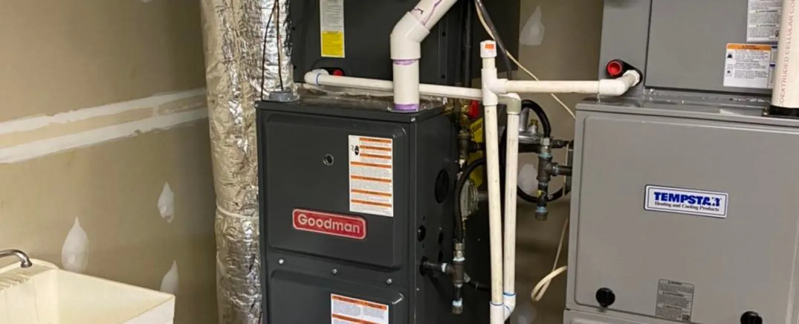 Basement HVAC system featuring Goodman and Tempstar furnaces with ductwork and plumbing beside a utility sink.