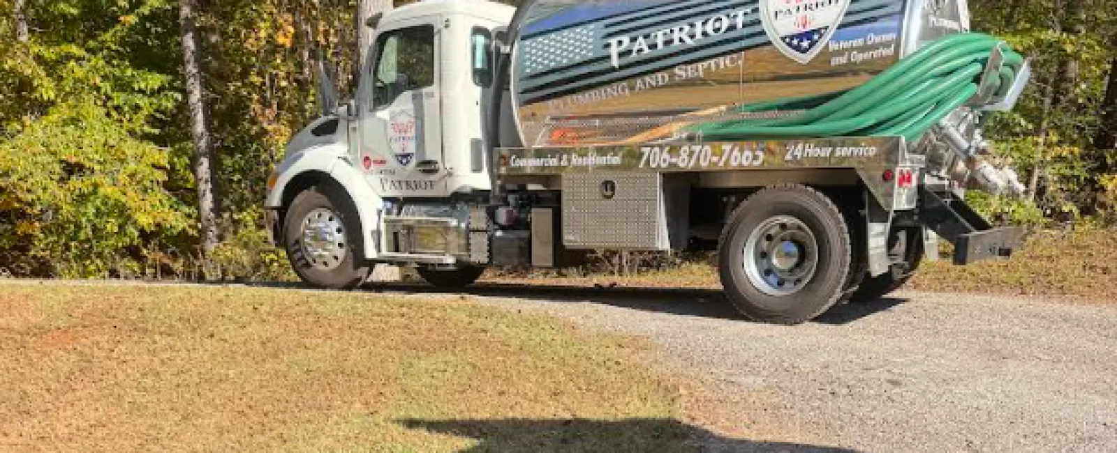 Patriot Plumbing and Septic service truck with hoses parked on a gravel driveway surrounded by trees in autumn.
