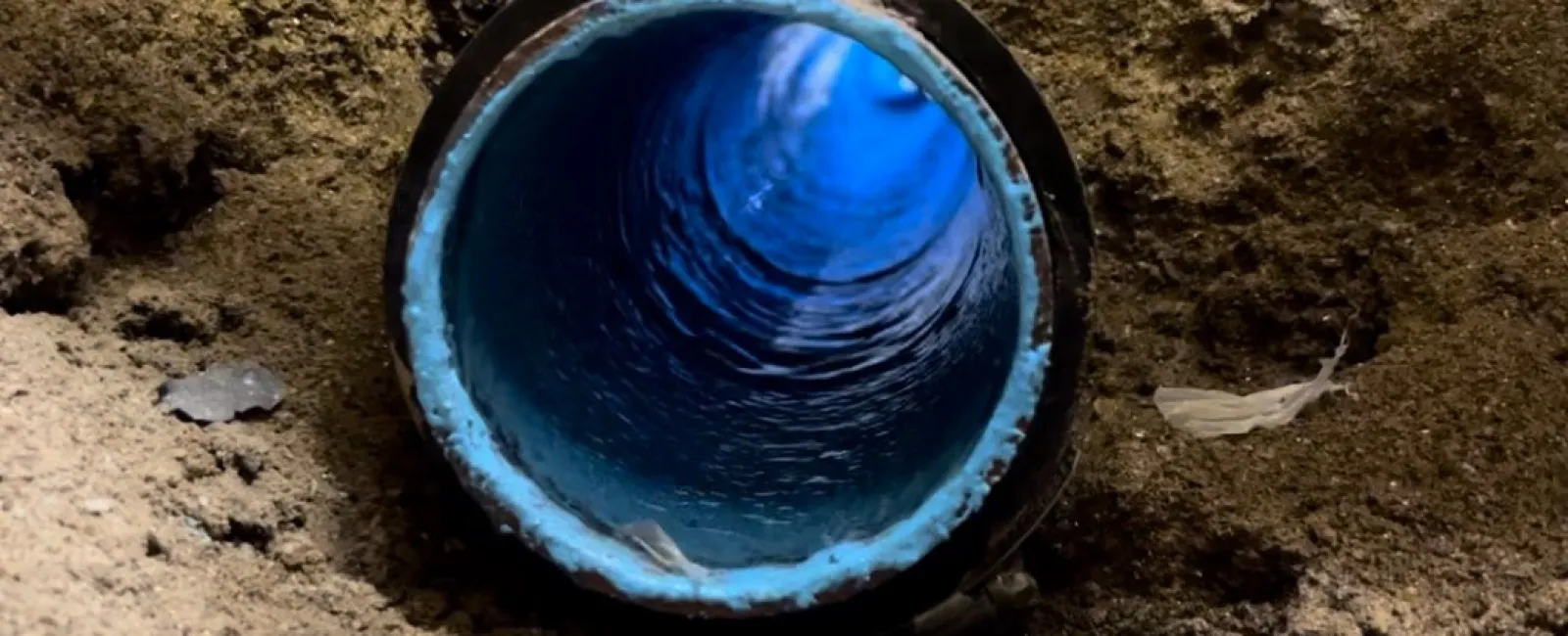 Close-up of a blue water pipe in soil with visible water flow inside surrounded by dirt and debris.