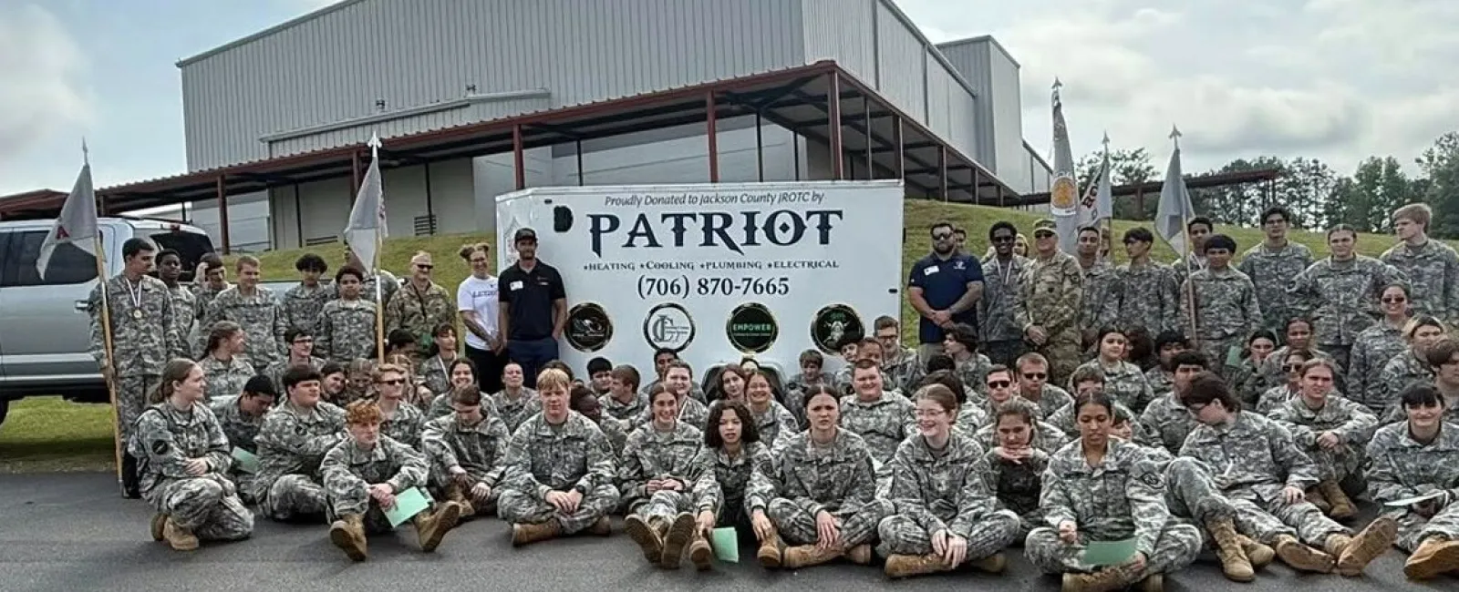 Group of uniformed soldiers posing in front of a Patriot sign and a white truck outside a large building on a cloudy day