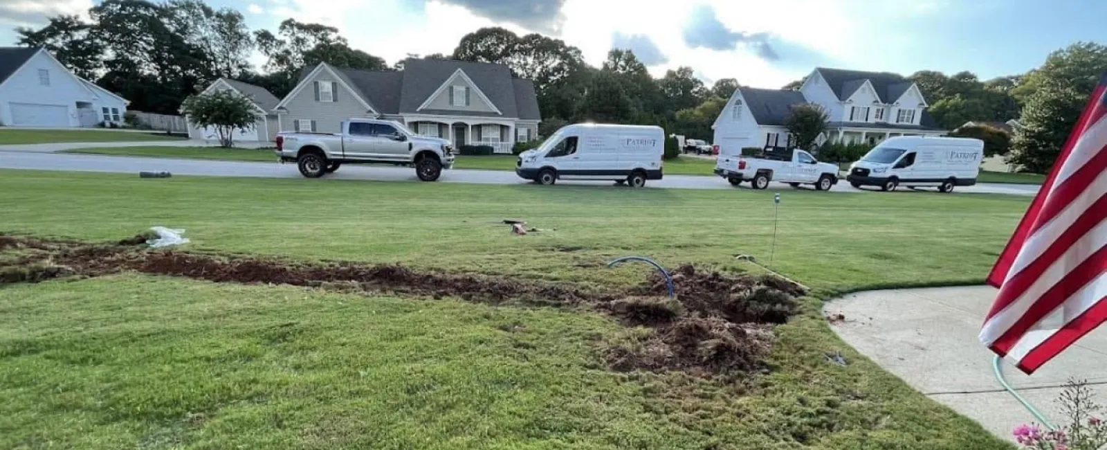 Residential lawn with trench dug for installation, parked service vans and trucks in background under partly cloudy sky.