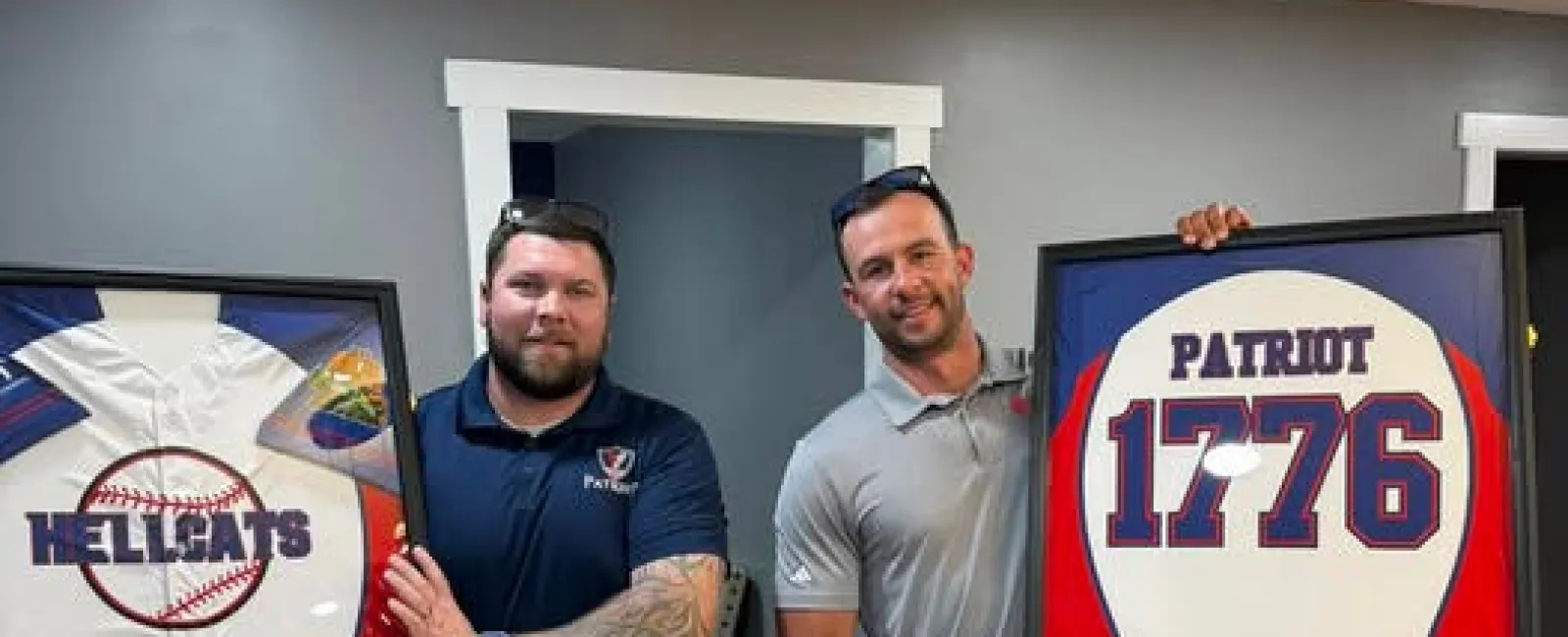 Two men holding framed baseball jerseys, one with Hellcats logo, the other with Patriot 1776 design inside a room.