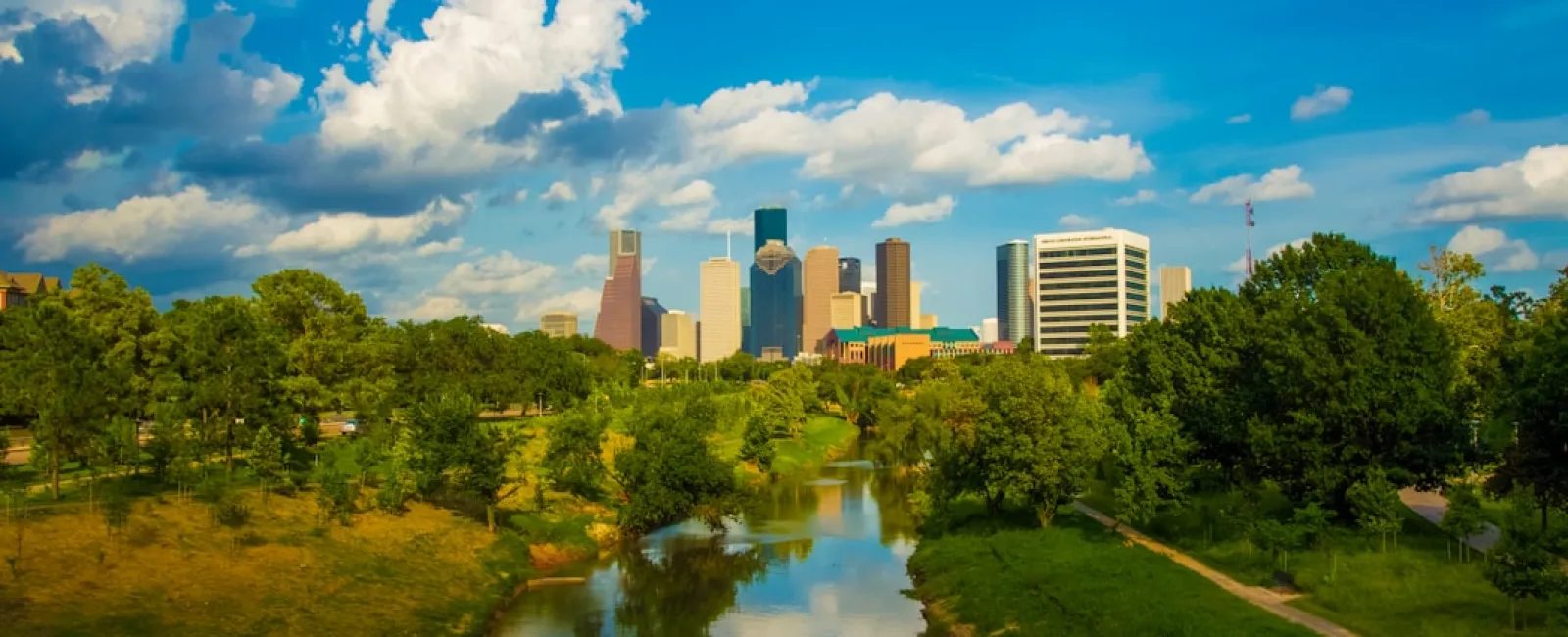 City skyline with tall buildings reflected in a calm river surrounded by green trees under a blue sky with clouds.