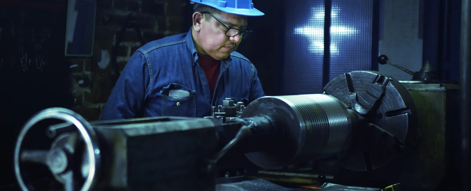 Man wearing blue hard hat operating industrial lathe machine in dimly lit workshop environment.
