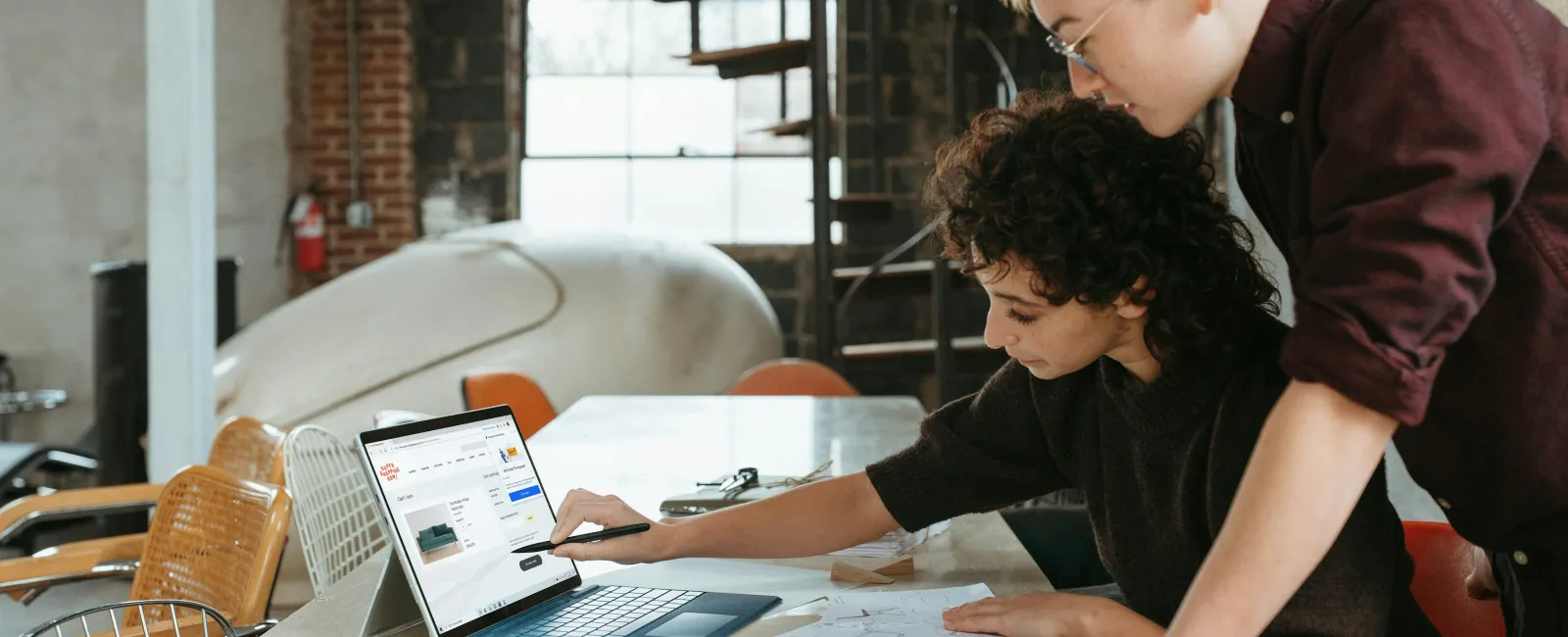 Two designers collaborate over a laptop in a modern workspace with sketches and furniture elements.