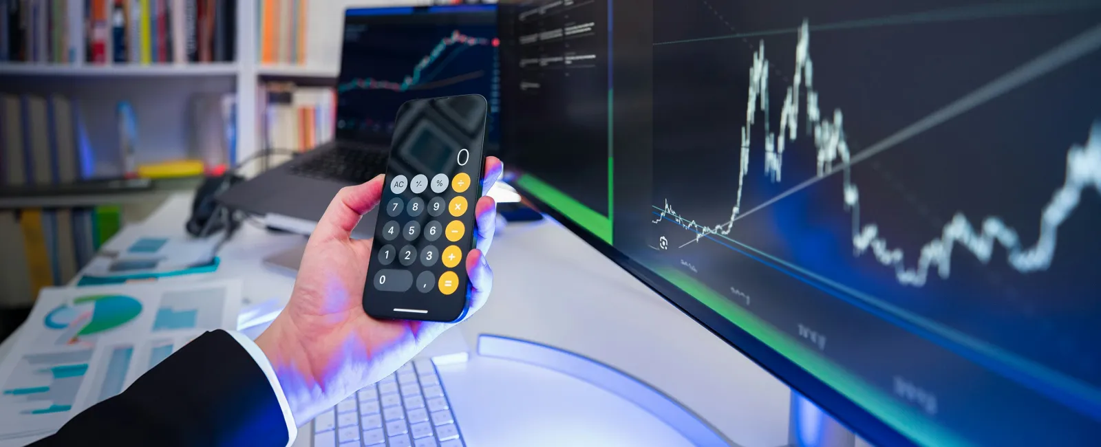 Businessperson holding smartphone with calculator app, analyzing stock market charts on dual computer monitors.