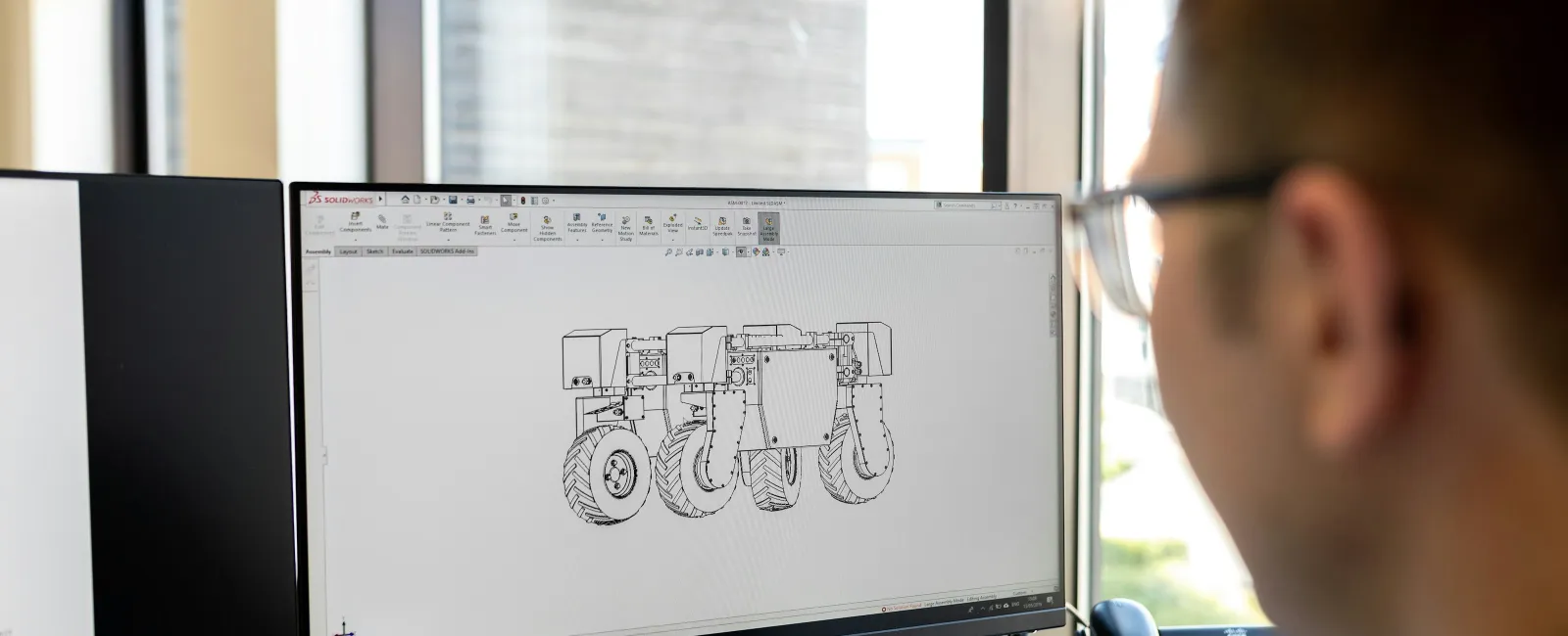 Engineer viewing a 3D CAD model of a vehicle with multiple wheels on a Dell monitor in a bright office.